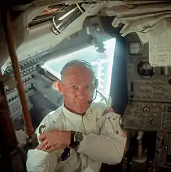 an astronaut with short hair and wearing a watch, flight suit and commset looks at the camera