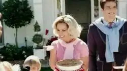 Image of a blond woman. She is a housewife and carries a pie in her left hand. Her hair is styled in 1950s fashion. She is wearing a pink polo shirt. Surrounding her are her husband and children.