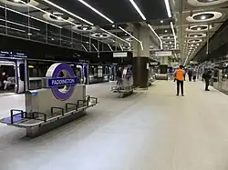 A wide island platform, with glass platform edge doors at either side. There are purple roundels labelled Paddington and benches in the centre. The ceiling is concrete with large circular light fixtures.