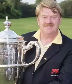 Portly caucasian man with light brown hair and moustache wearing a blazer atop polo shirt with the Walker Cup logo and holding a trophy cup